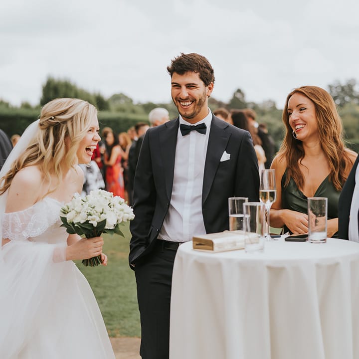 Bride holding white rose wedding bouquet socialises with guests outside around white circle tables 