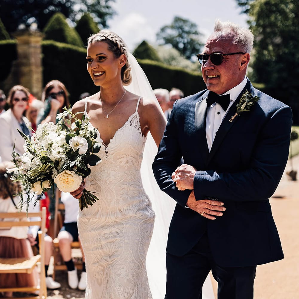 Father of the bride in sunglasses and black tie walks the bride in lace wedding dress down the aisle for outdoor wedding ceremony 