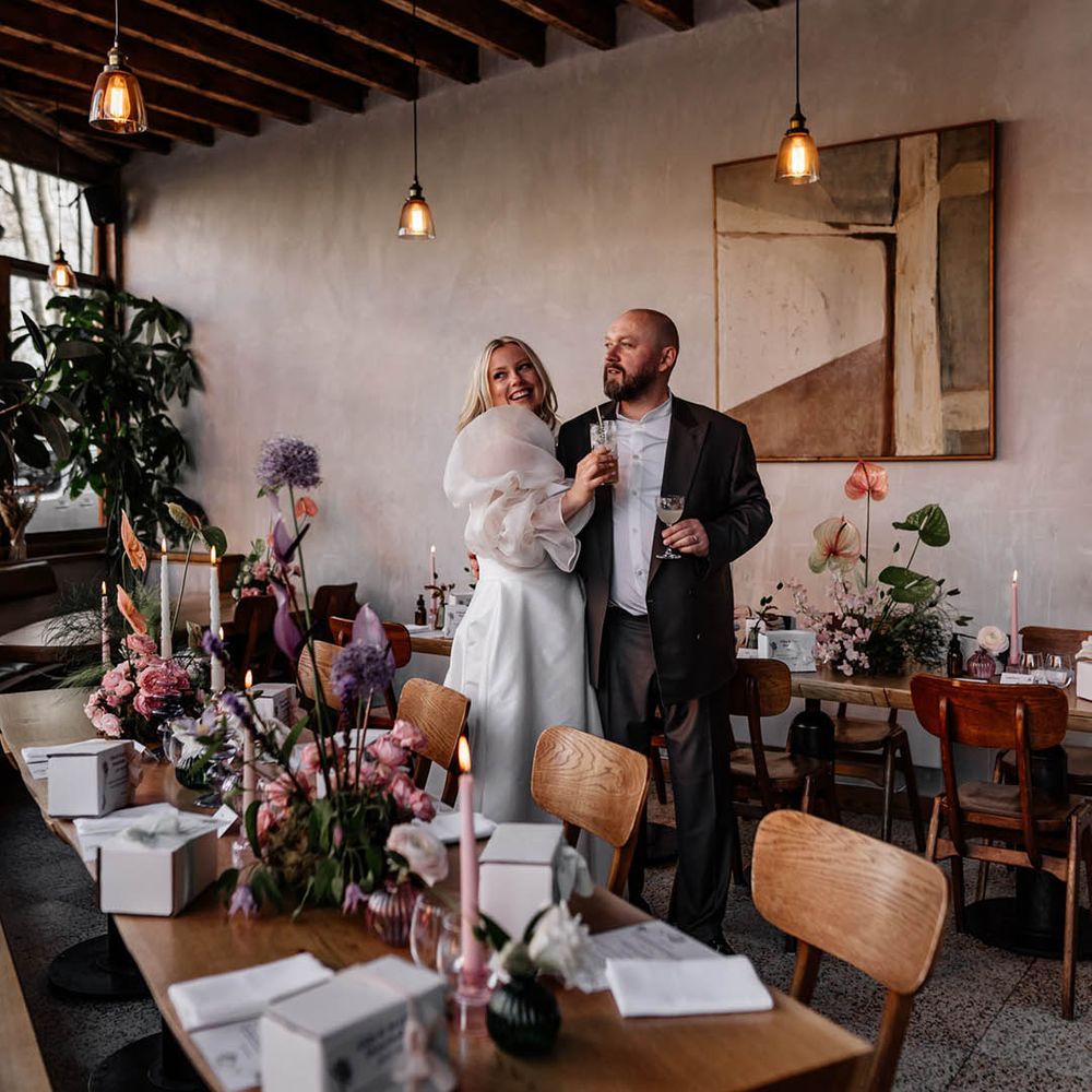 Bride and groom standing in the middle of their wedding reception with tall purple allium stems, pink anthuriums and taper candle table decor 