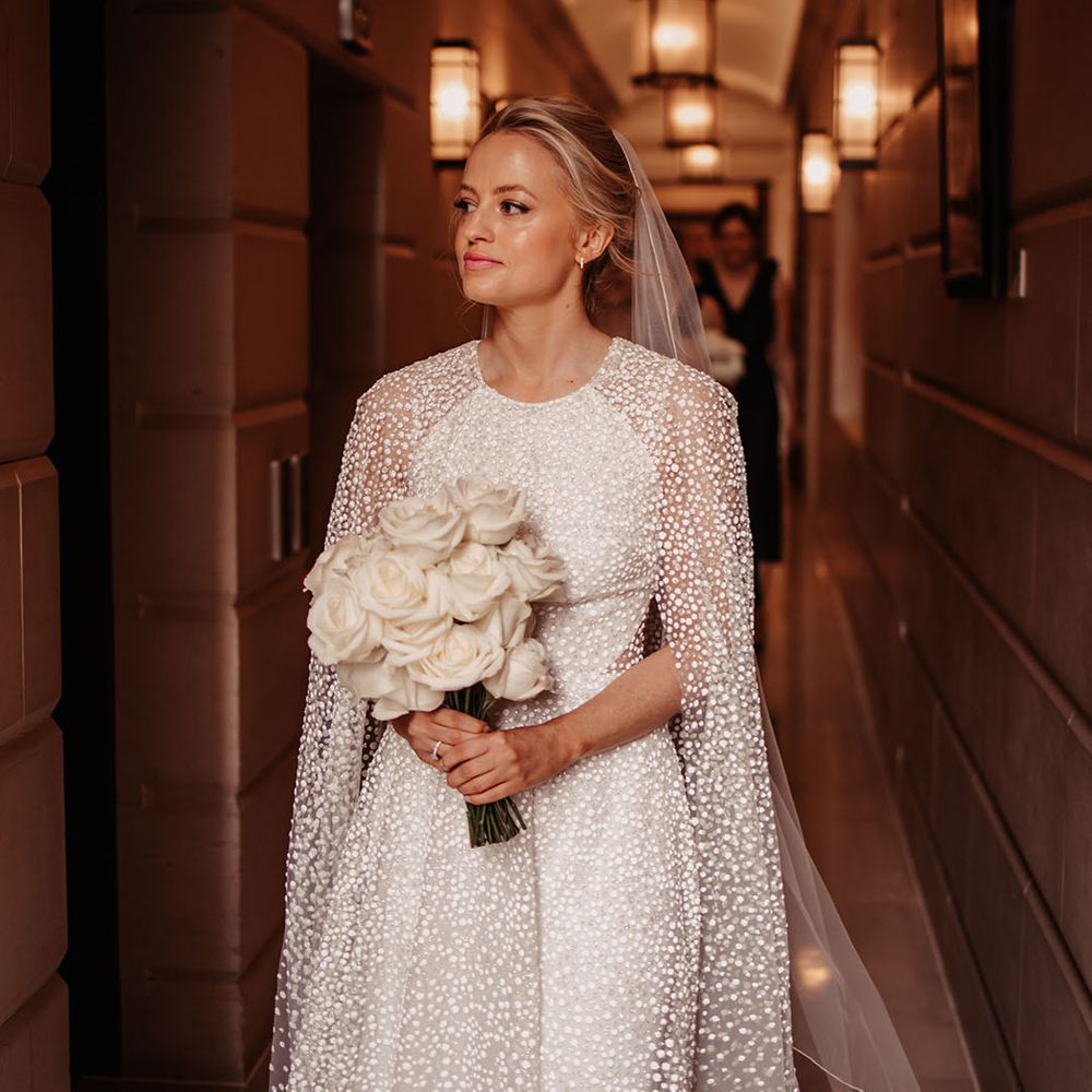 Blonde bride walks with Jesus Peiro cape and wedding dress with a classic white rose bouquet 
