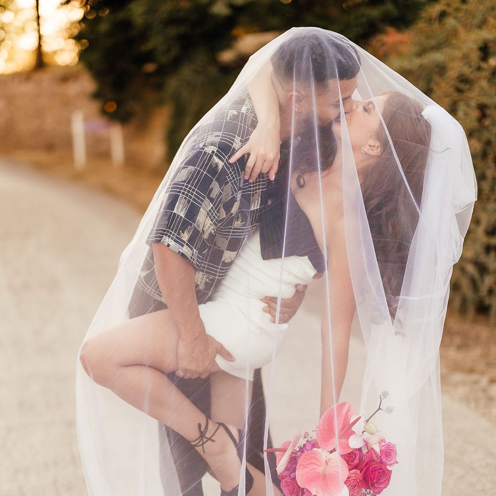 Bride wears white mini dress and holds brightly coloured bouquet as she kisses her groom beneath veil