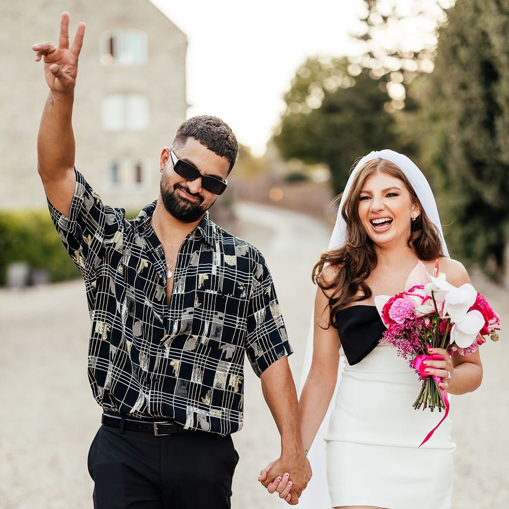 Groom wears open shirt and sunglasses whilst holding his brides hand who wears mini wedding dress 