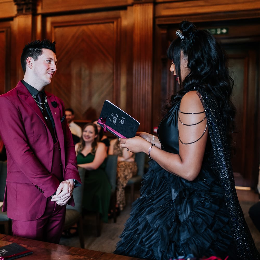Groom wears wine red suit with chain detailing to shirt as he listens to his bride give personal wedding vows 