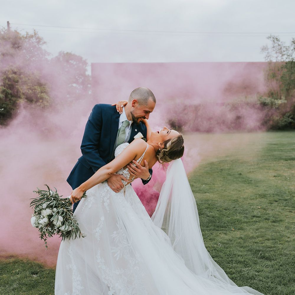 Groom in a dark blue suit, grey waistcoat and a sage green tie leans the bride back as pink smoke goes off in the background