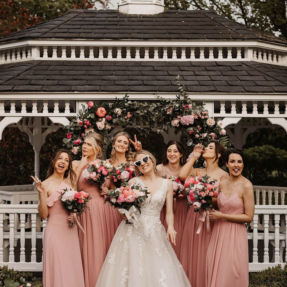 Bridesmaids in pink dresses do funny poses with the bride in a lace wedding dress and cat eye retro sunglasses 