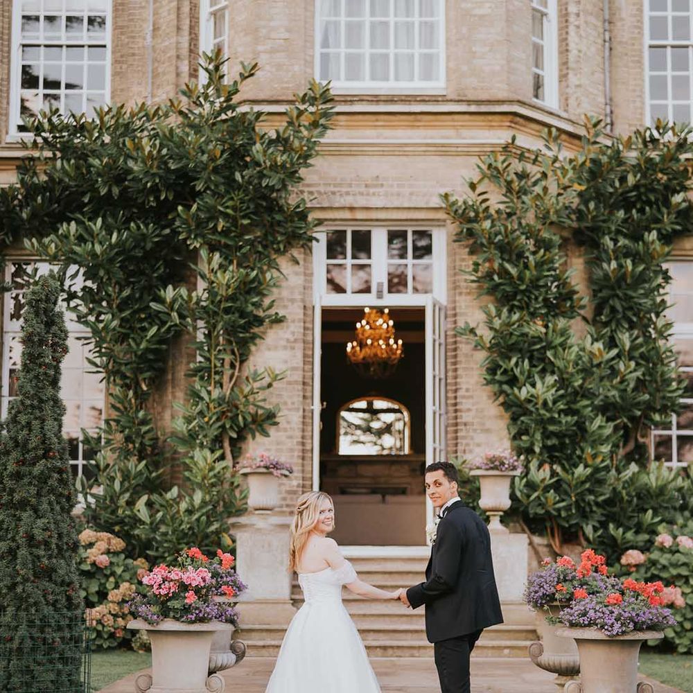 Bride in off shoulder wedding dress with lace detail and groom in black tux holding hands outside Hedsor House