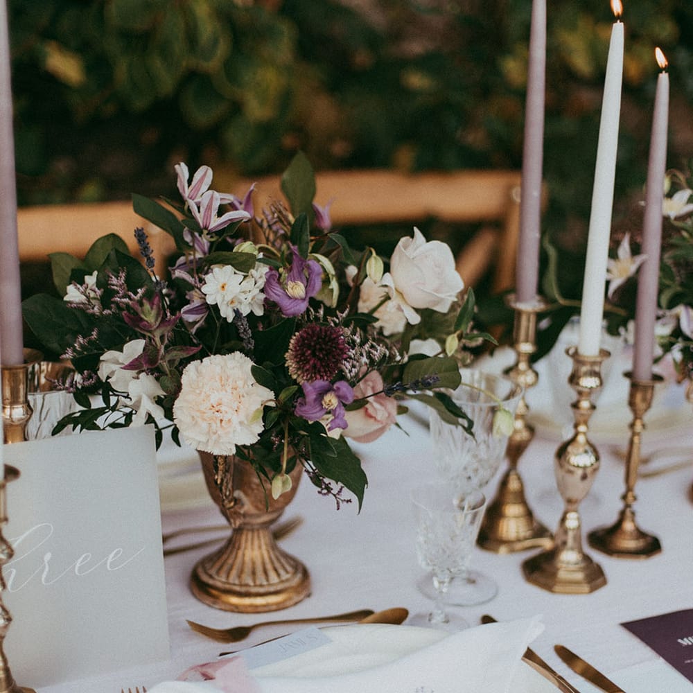 Floral arrangement with white carnations, purple tulips, roses and foliage in gold vase, white and lavender tapered candles in gold candle holders, and gold cutlery
