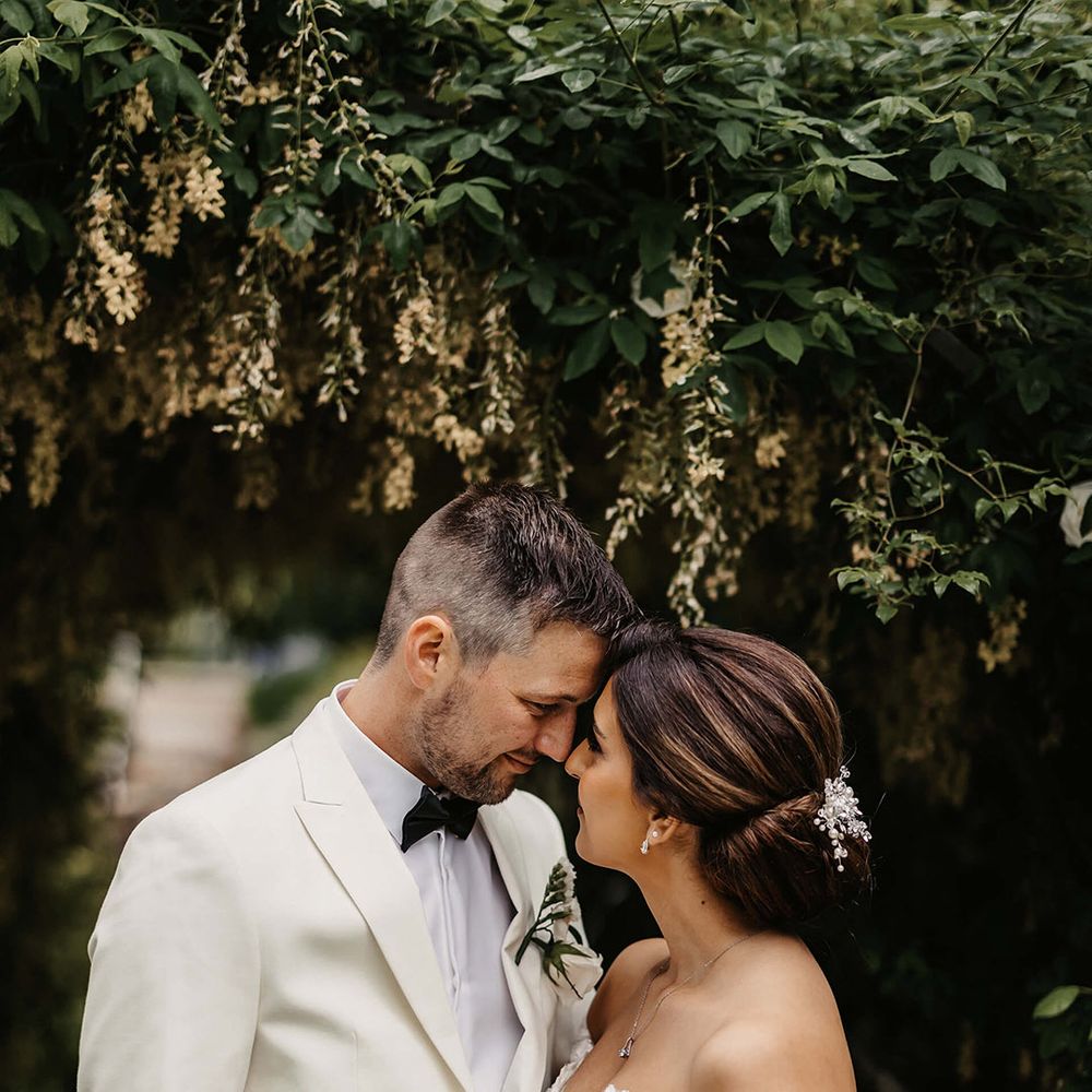 The bride wears an off the shoulder lace wedding dress smiling up at the groom in a white tuxedo 