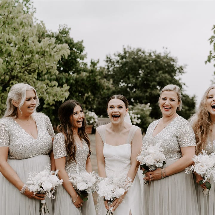 Bride laughs with her bridesmaids in pre loved silver sequin dresses holding white dried flower bouquets 