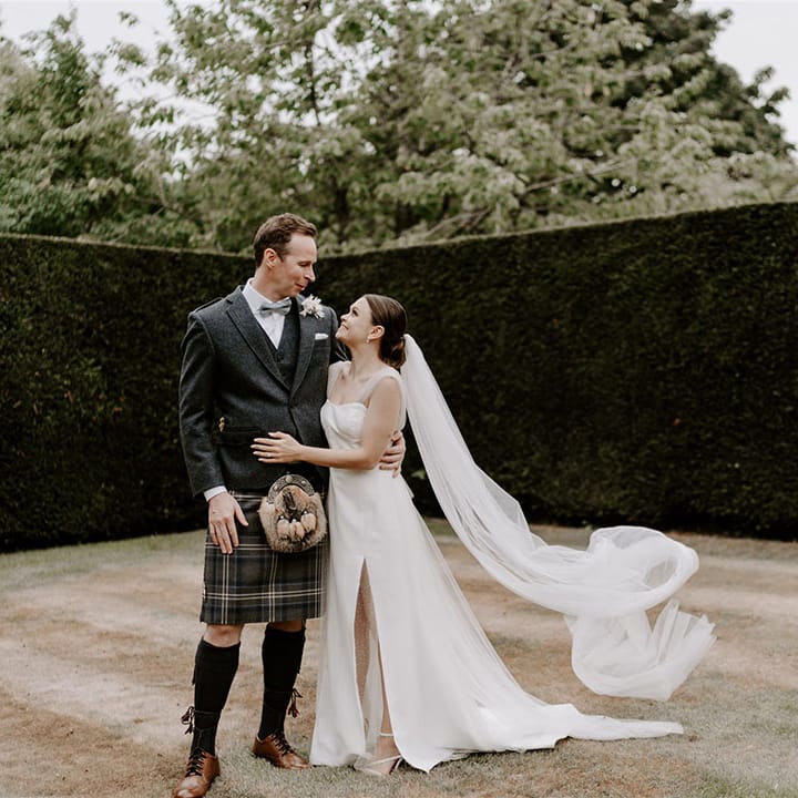 The groom in a kilt and matching tweed waistcoat an d blazer with blue bow tie stands with the bride in a sparkly wedding dress and cathedral length veil 