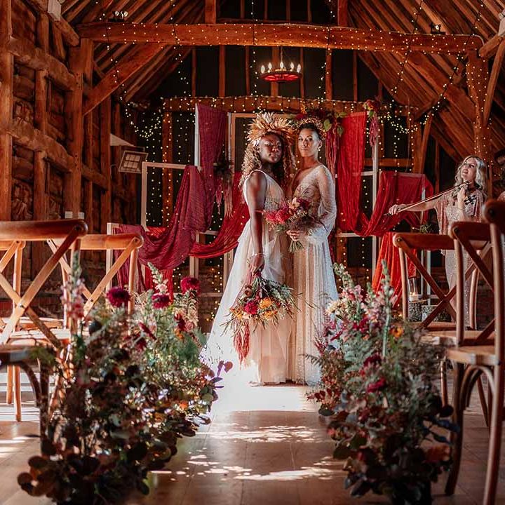 Brides in complimenting boho wedding dresses with embroidered stars and polka dots and gold bridal crowns kissing in front of large orange smoke bomb at Silchester Farm