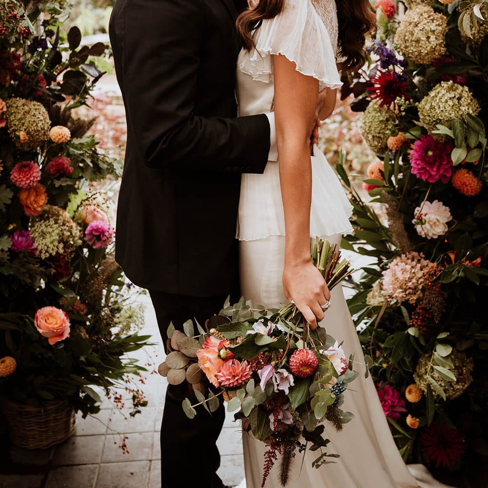 Groom in classic black tuxedo with hand painted silk cravat and handkerchief kissing bride in Bride in v neck satin wedding dress with sheer butterfly sleeves and ruffled layer on top standing in large floral arch with baby's breath, pink and white carnations, garden roses and dried flowers