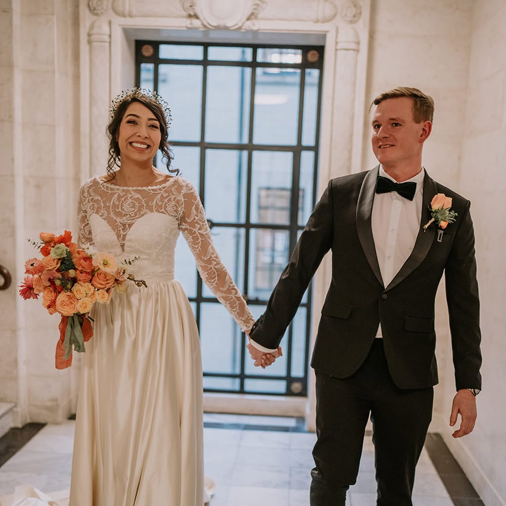 Groom in stylish black tuxedo with black bowtie and light orange rose boutonniere holding hands with bride in  taffeta material princess wedding dress with long lace sleeves 