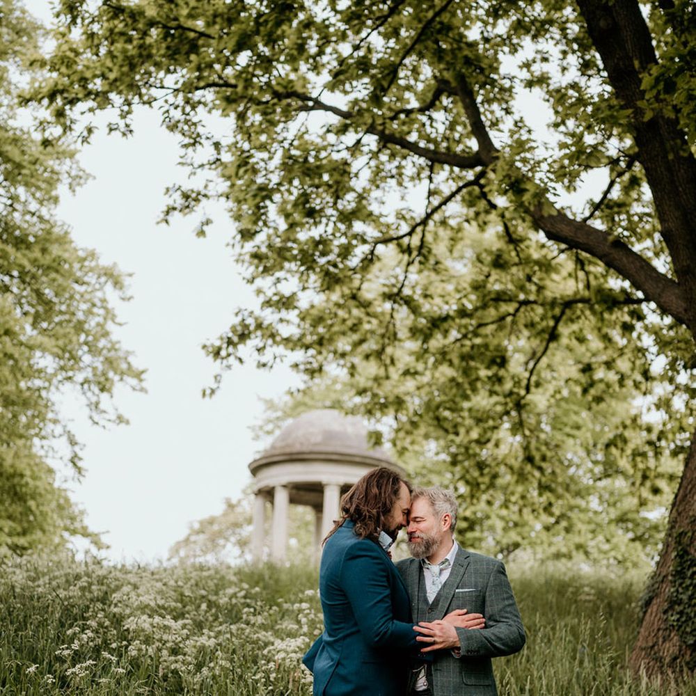 Groom in blue suit and groom in tartan suit embrace outdoors surrounded by lush greenery for botanical wedding 