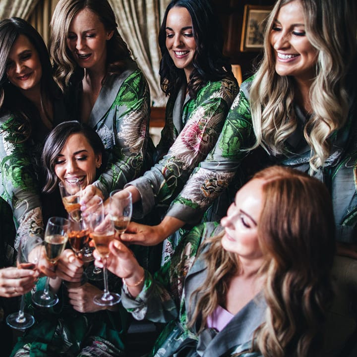 Bridesmaids in grey patterned robes with flower print cheers each other and the bride as they get ready on the morning of the wedding 