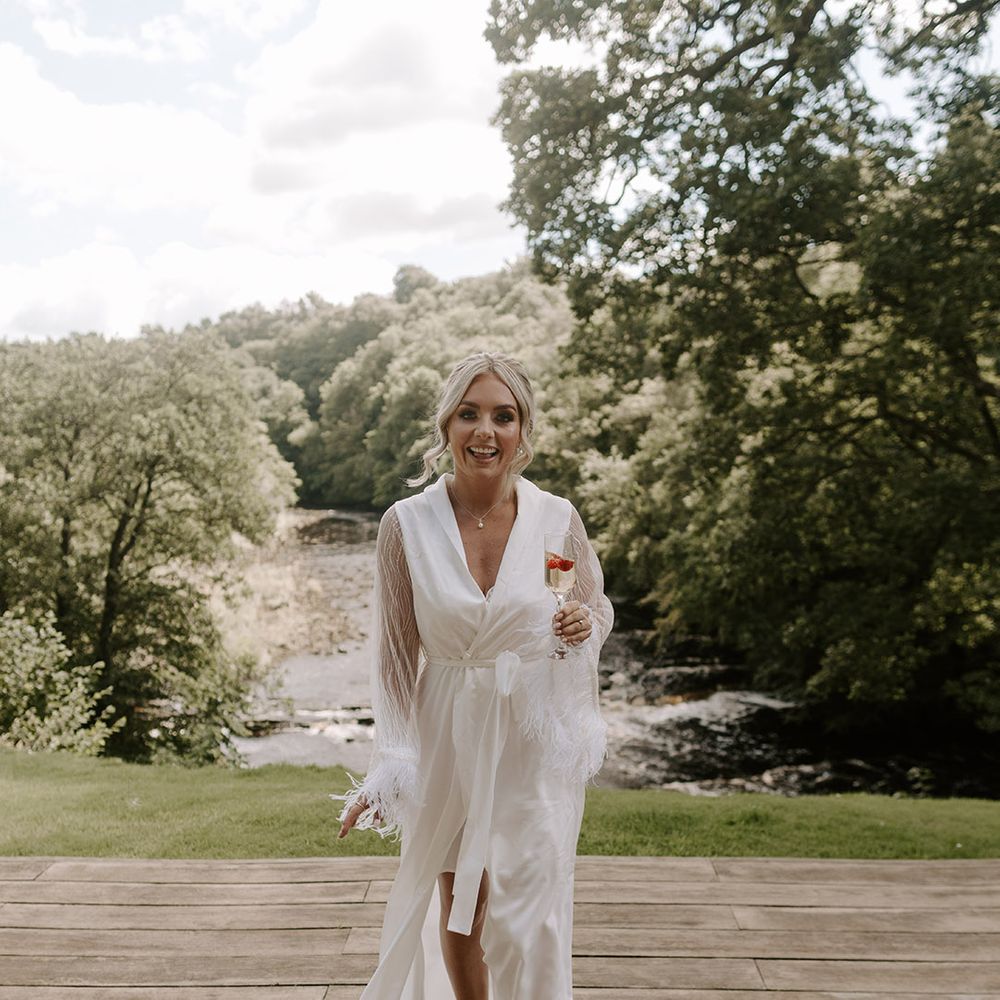 The bride wears a long white robe with sheer sleeves and feather cuffs in white pearl flip flops drinking champagne 