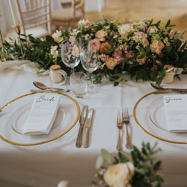 Gold rim clear charger plates with black and white wedding menu stationery on a white tablecloth with pastel wedding flower centrepiece 