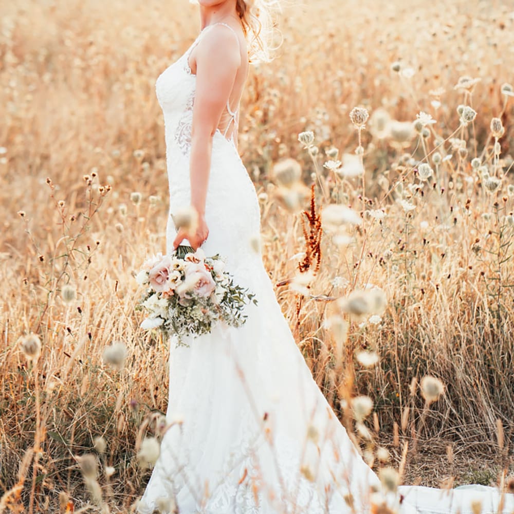 Smiling bride with her blonde hair in a half up half down hairstyle in a fitted lace wedding dress with low back holding a white bouquet 