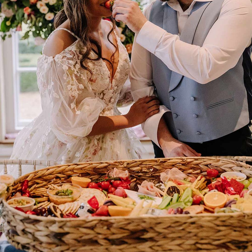 Groom in cornflower blue waistcoat and black suit trousers giving bride in v-neck long sleeve shoulder cut out 3d appliqué wedding dress with flecks of gold some wedding grazing platter 