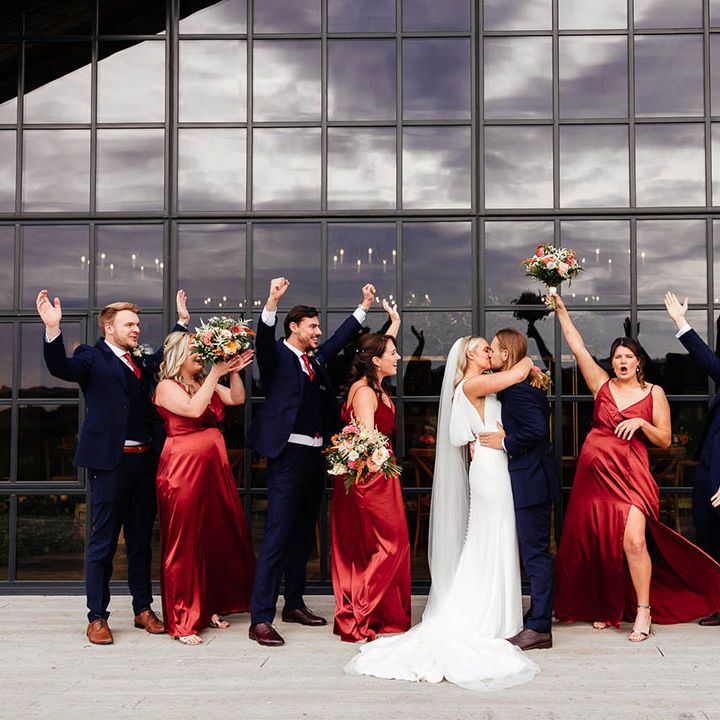 The bridesmaids in red satin bridesmaid dresses and the groomsmen in blue suits celebrate together as the bride and groom kiss