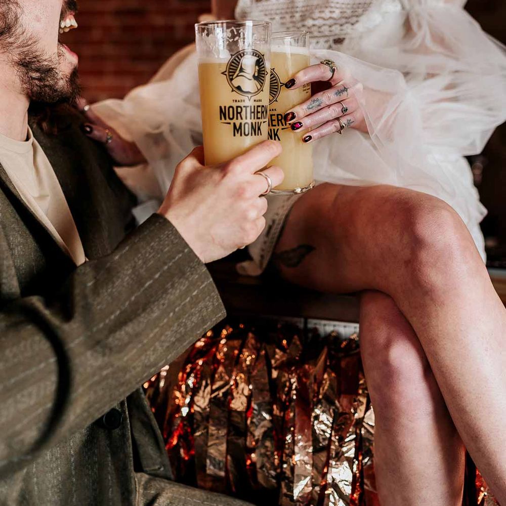 Bride in 70s inspired boatneck short lace wedding dress with detachable tulle puff sleeves cheersing beer with groom in deep brown wedding suit and off white t shirt 