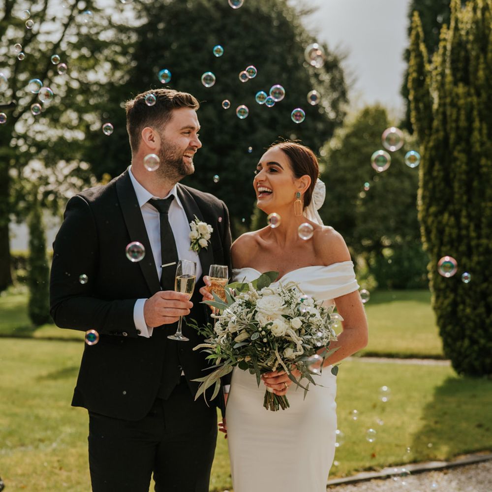 Bride in off the shoulder wedding dress with the groom in a black tuxedo drinking a glass of champagne surrounded by bubbles 