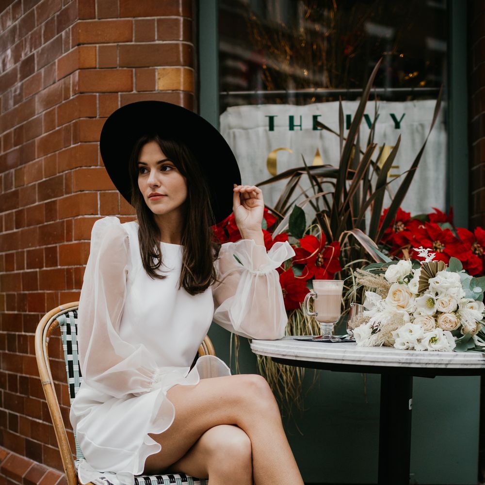 Bride sitting at a cafe in knee length wedding dress and fedora hat 