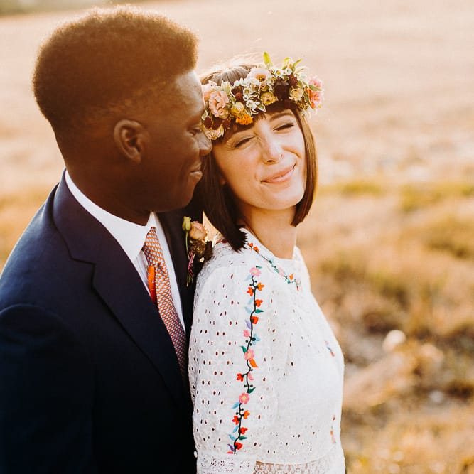 Golden hour portraits with groom in a navy suit smiling at his bride in a colourful flower crown 