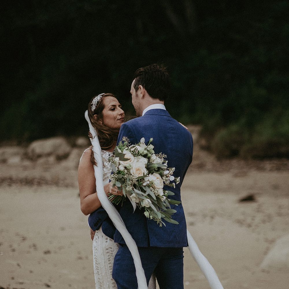 Bride & groom stand together on the beach as brides veil blows around them