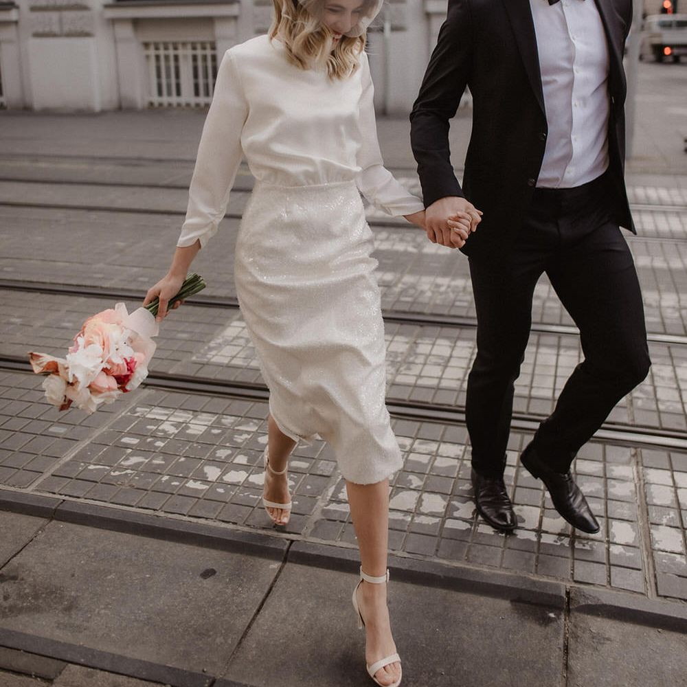 Bride and groom walking through the city, bride is wearing short mesh veil over her face with white, red and pink flowers on her head, and a long sleeve mid length wedding dress with sequin pencil skirt