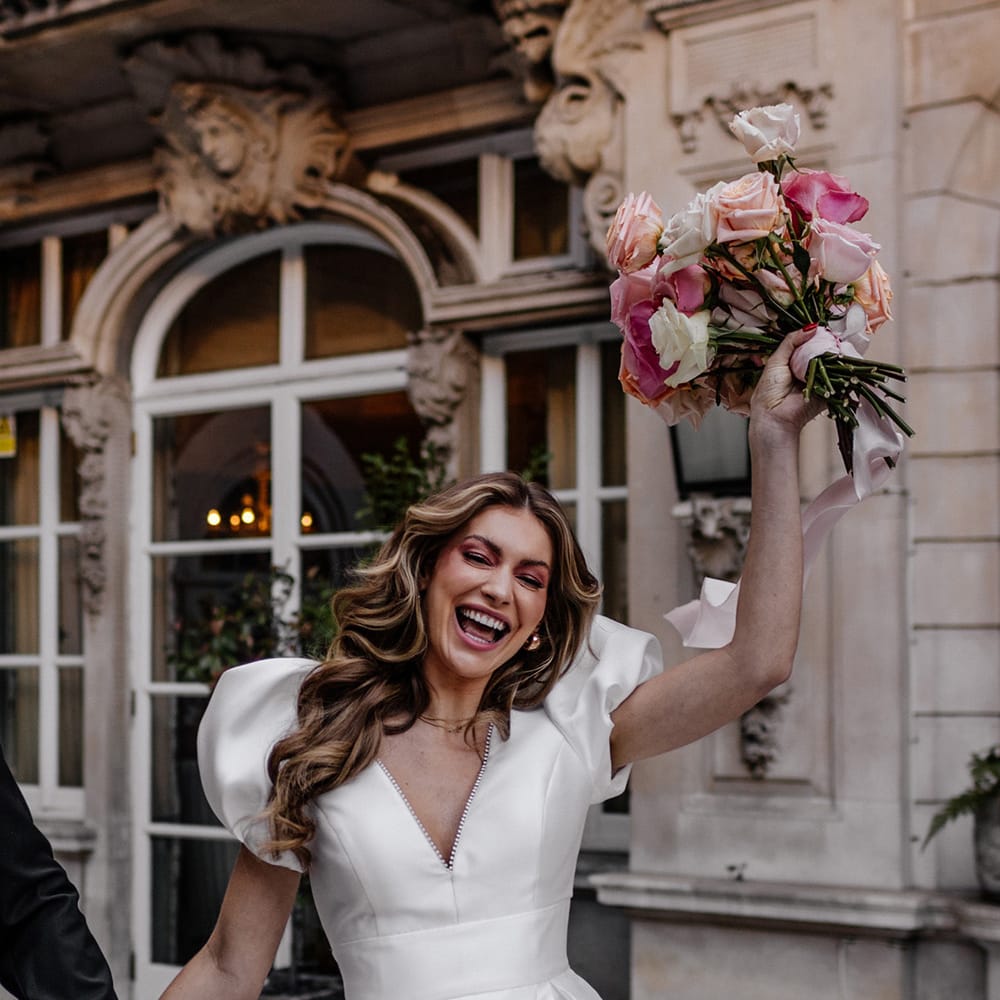 happy bride with long wavy hair, and pink eye shadow waving her blush pink, peeled back rose wedding bouquet in the air 
