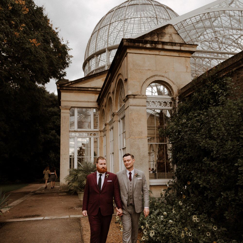 Groom in a burgundy suit holding hands with his husband in a beige check suit outside Syon Park wedding venue 