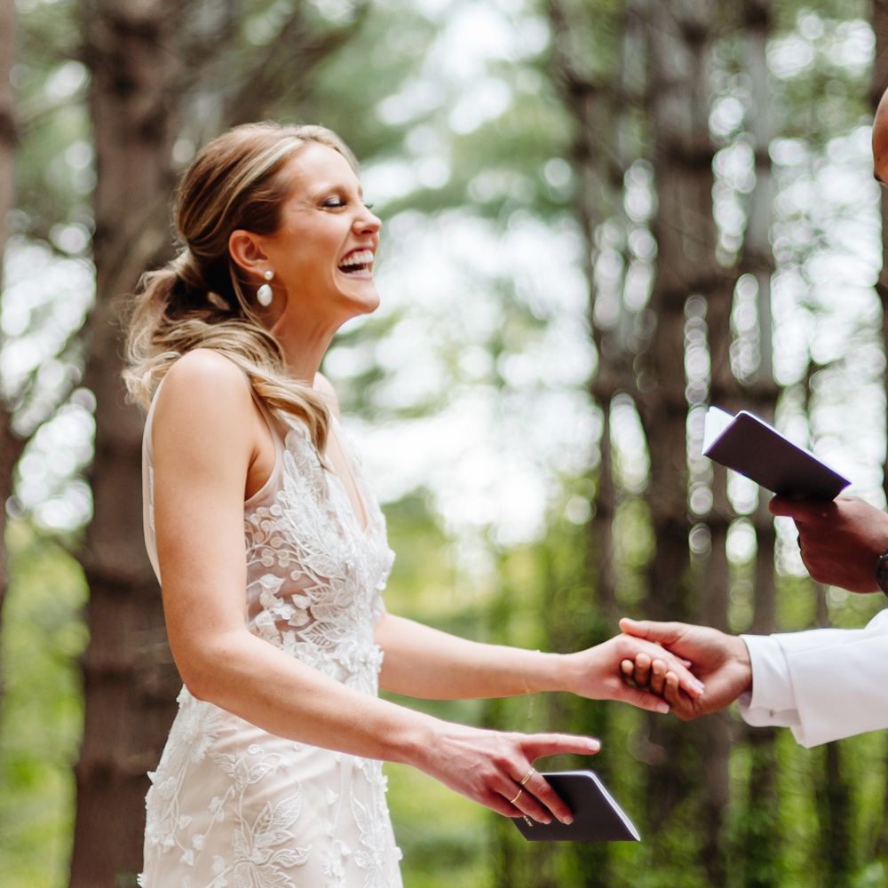 Smiling bride in white embroidered Made With Love wedding dress holds hands with groom in white tuxedo jacket in woodland as they read private vows to one another after backyard wedding