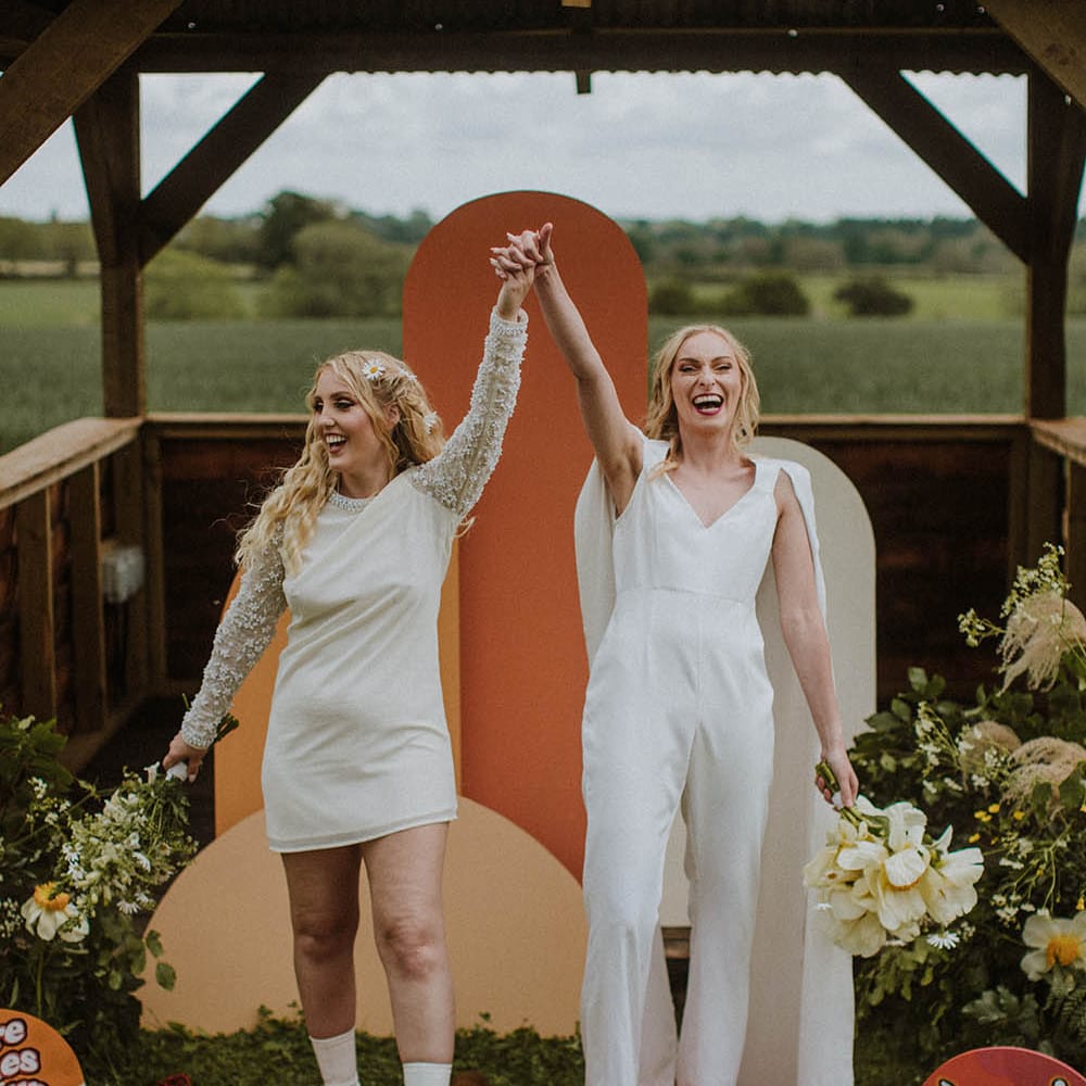 Sixties wedding with two brides in a mini wedding dress and bridal jumpsuit holding hands descending up the aisle 