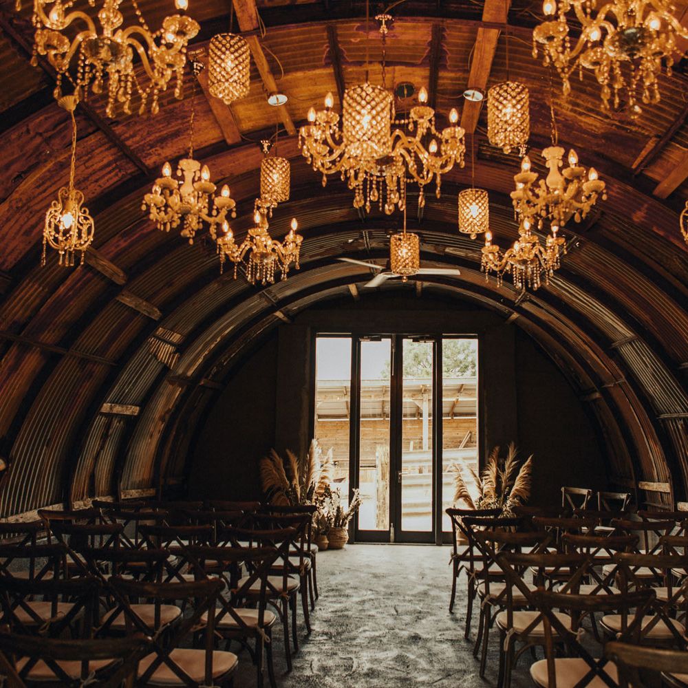 Hut with corrugated iron semi circular roof and assorted lit chandeliers hanging from the roof over chairs set out for wedding ceremony at Anran Devon