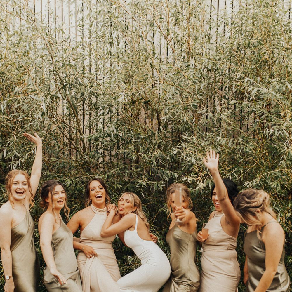Bride in white Made With Love wedding dress stands outside with bridesmaids in satin sage green bridesmaids dresses and high necked beige wedding dresses as they all laugh at Anran Devon