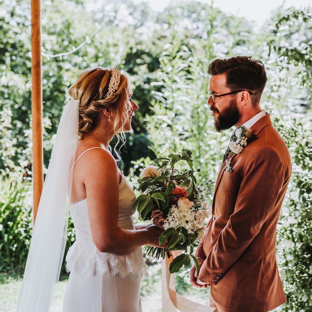 Bride & groom look lovingly at one another in front of green foliage background under tipi