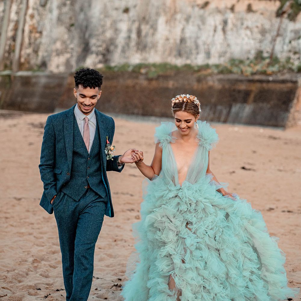 Groom in a blue wool suit holding hands with his bride in a layered tulle mint green wedding dress at their beach elopement 