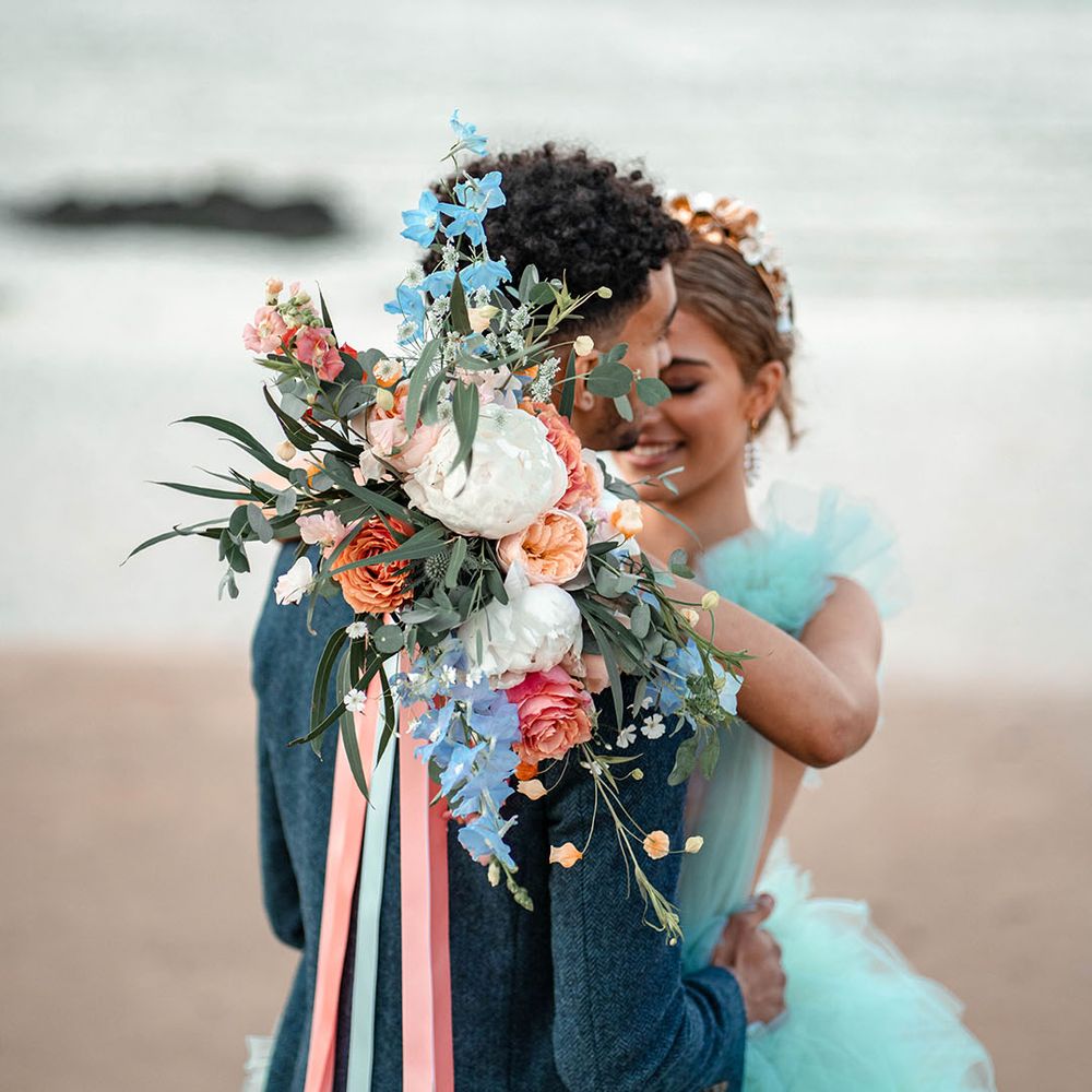 Bride holding her pastel bouquet tied with ribbon over her grooms shoulder with white peonies, coral and peach roses and blue wild flowers