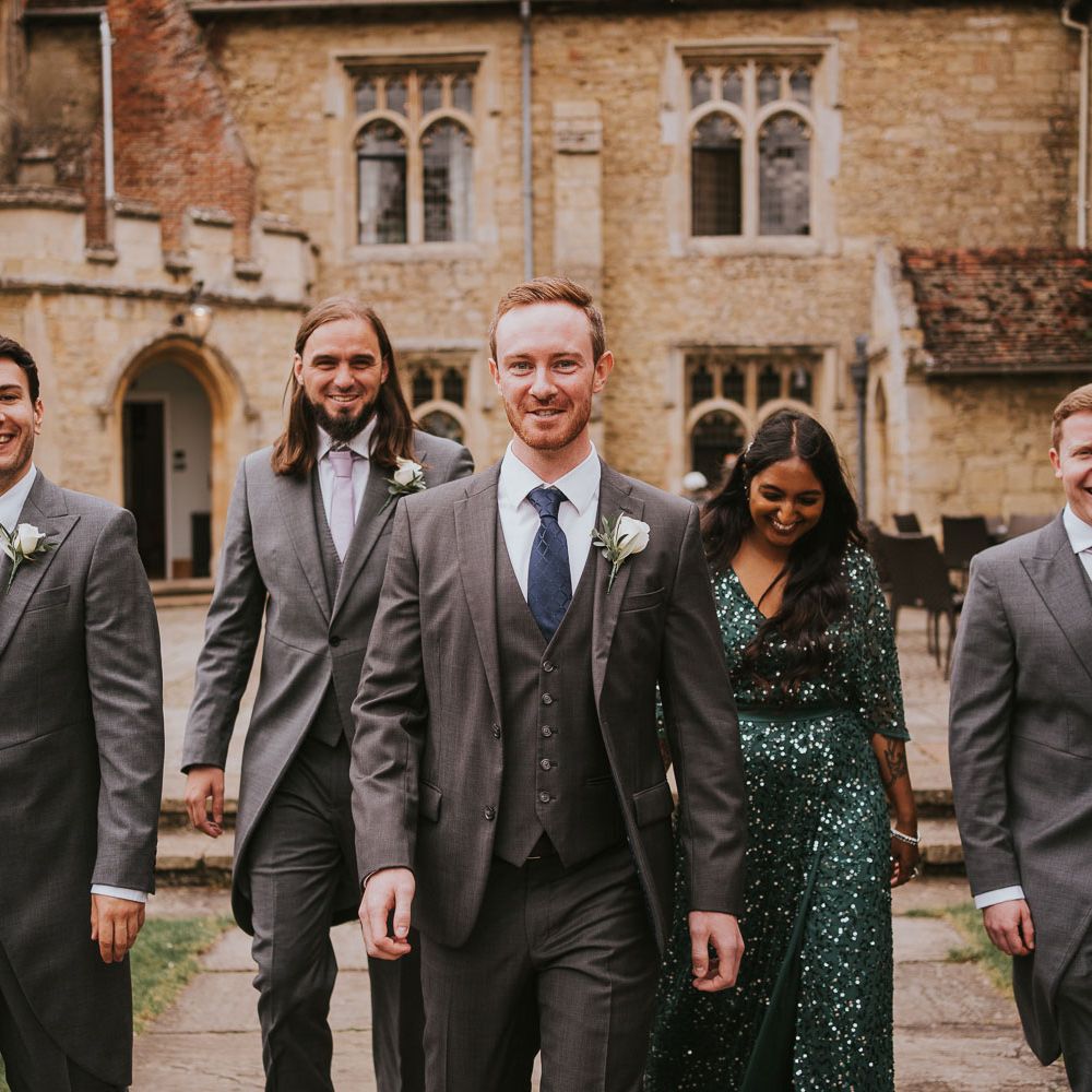 Groom in grey three piece suit with white rose buttonhole walks with three groomsmen in grey morning suits and woman in emerald sequin dress in the grounds of Notley Abbey