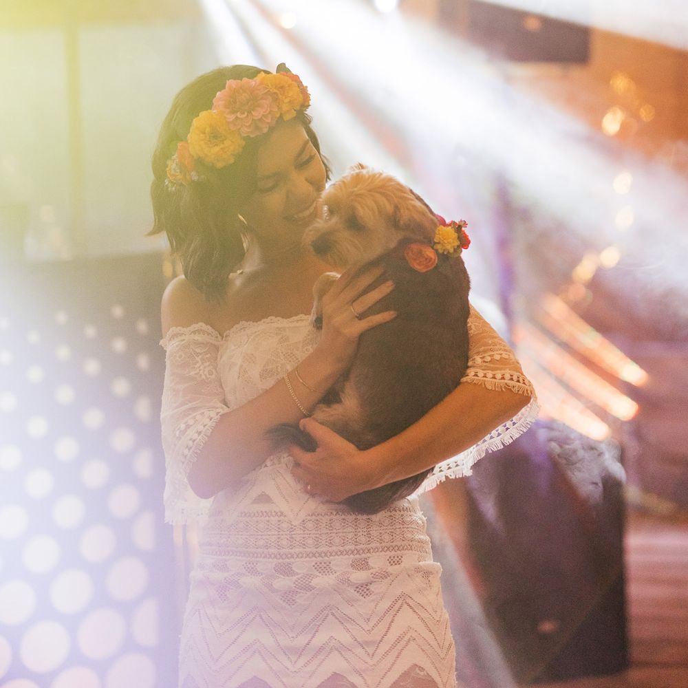 Bride in off the shoulder Grace Loves Lace wedding dress and flower crown holds dog in floral dog collar inside barn for evening wedding reception