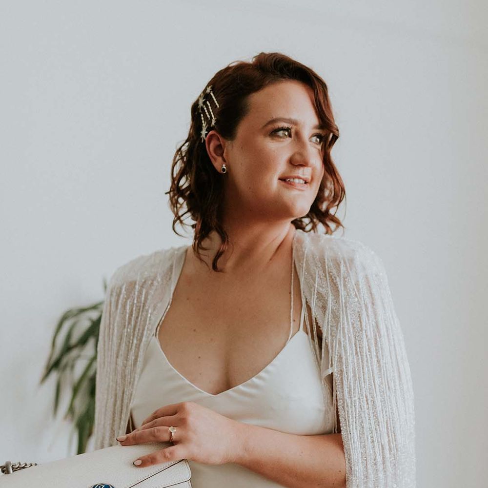 Bride wears her short hair slightly curled whilst holding white bag and wearing tassel Halfpenny London wedding cape