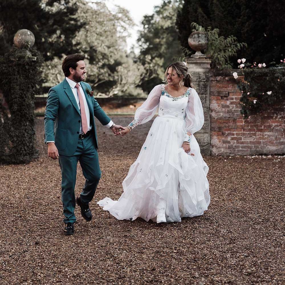 Groom in blue suit and pink tie holding hands with bride in long mesh sleeve wedding dress walks across drive at Iscoyd Park
