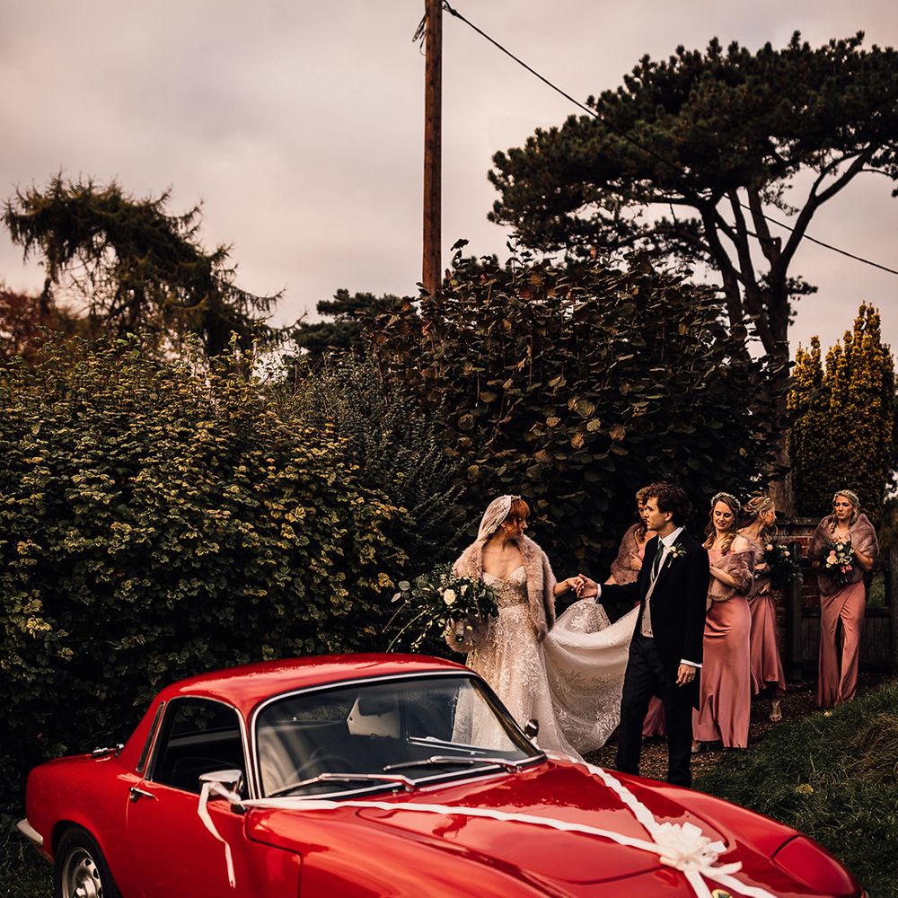 Bridesmaids help the bride to the vintage red car at Oxnead Hall wedding
