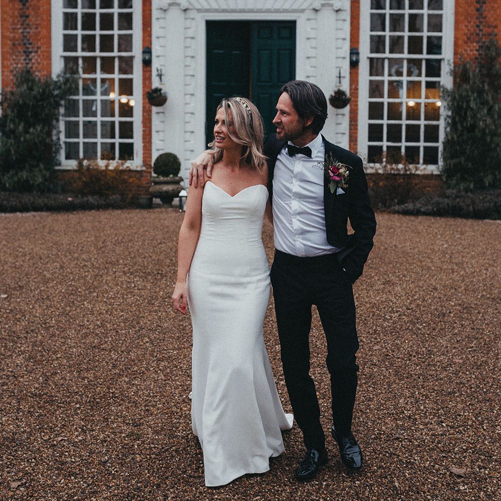 Groom wraps his arm around shoulders of his bride as they stand outdoors in front of Gosfield Hall 