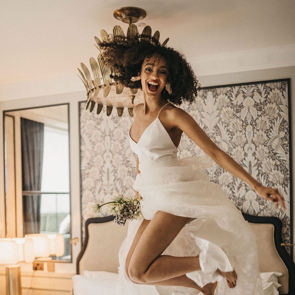 Black bride with naturally curly hair jumping on the bed at Mayfair Townhouse in a spaghetti strap wedding dress with lace skirt 