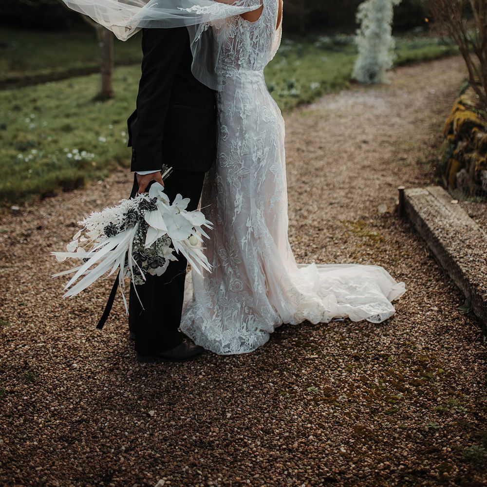 Bride in a lace wedding dress with watteau train embracing her partner in a black suit as he/him holds a winter wedding bouquet 