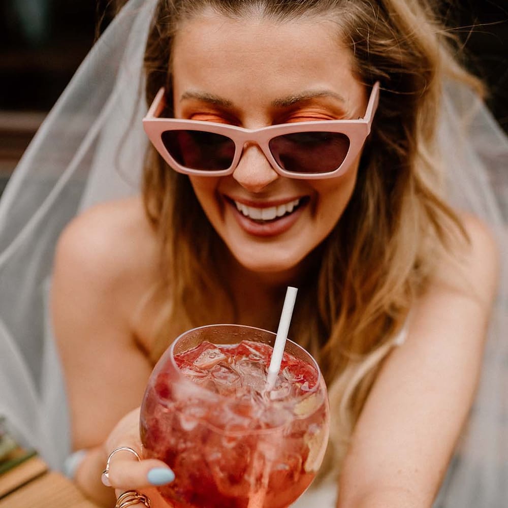 Bride in a retro pink sunglasses holding a ballon glass filled with a fruity cocktail, ice and a straw 