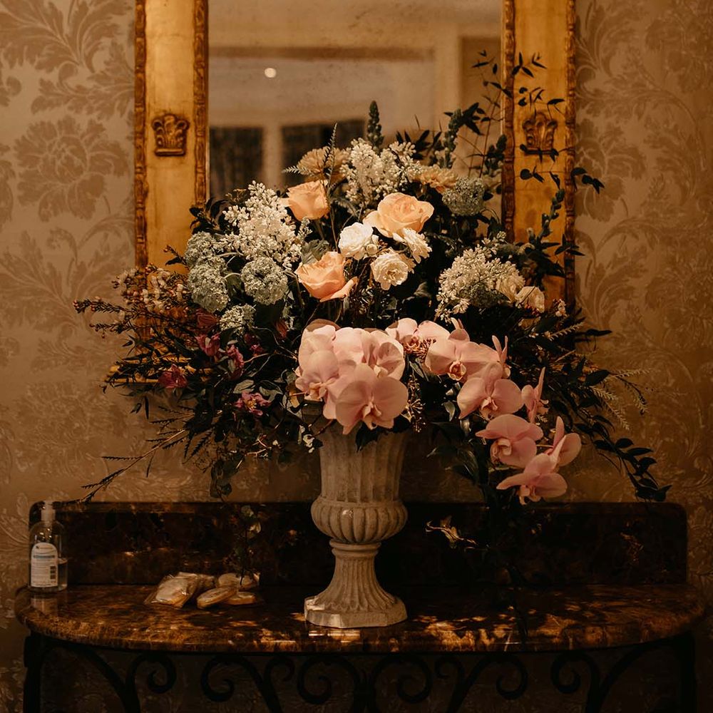 Large mixed neutral coloured flower display in cream ceramic vase behind large golden mirror
