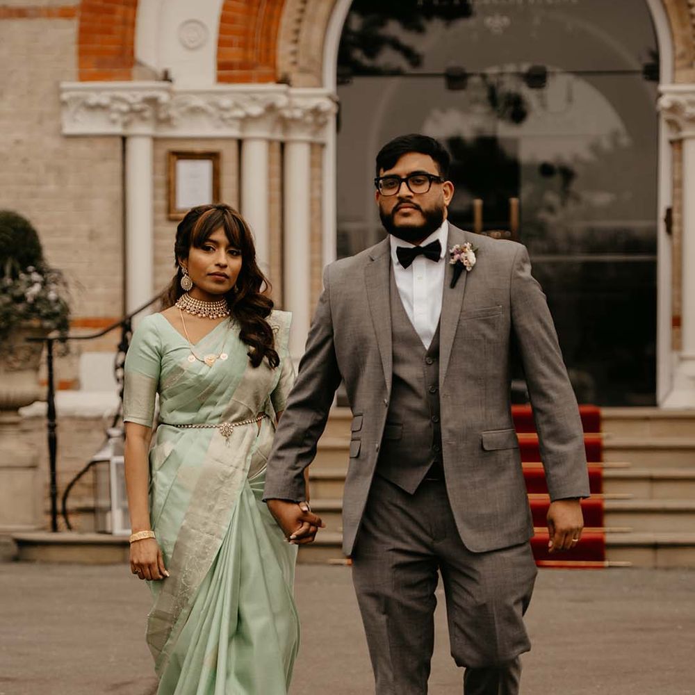 Bride in mint green saree with golden jewellery and groom in grey suit with black bow tie, holding hands